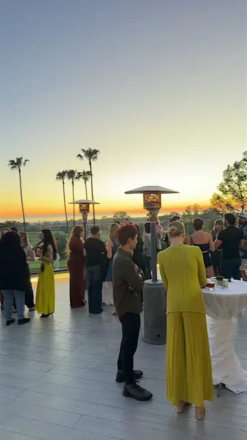 Attendees mingle on a terrace at sunset, with palm trees and the ocean on the horizon